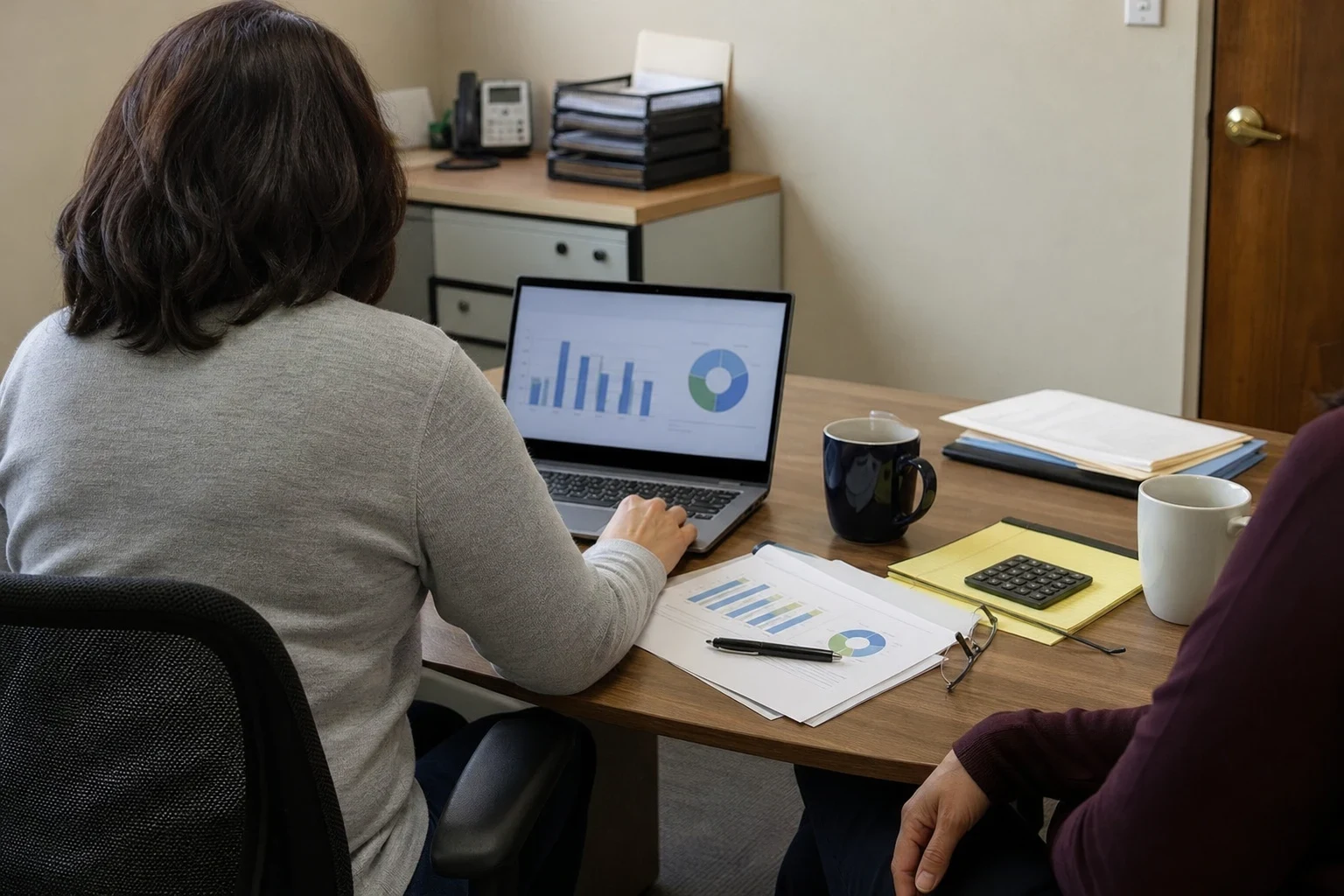 Woman at a small office desk reviewing bar and pie charts on a laptop with a client, printed reports, calculator, yellow notepad and coffee mugs on the table