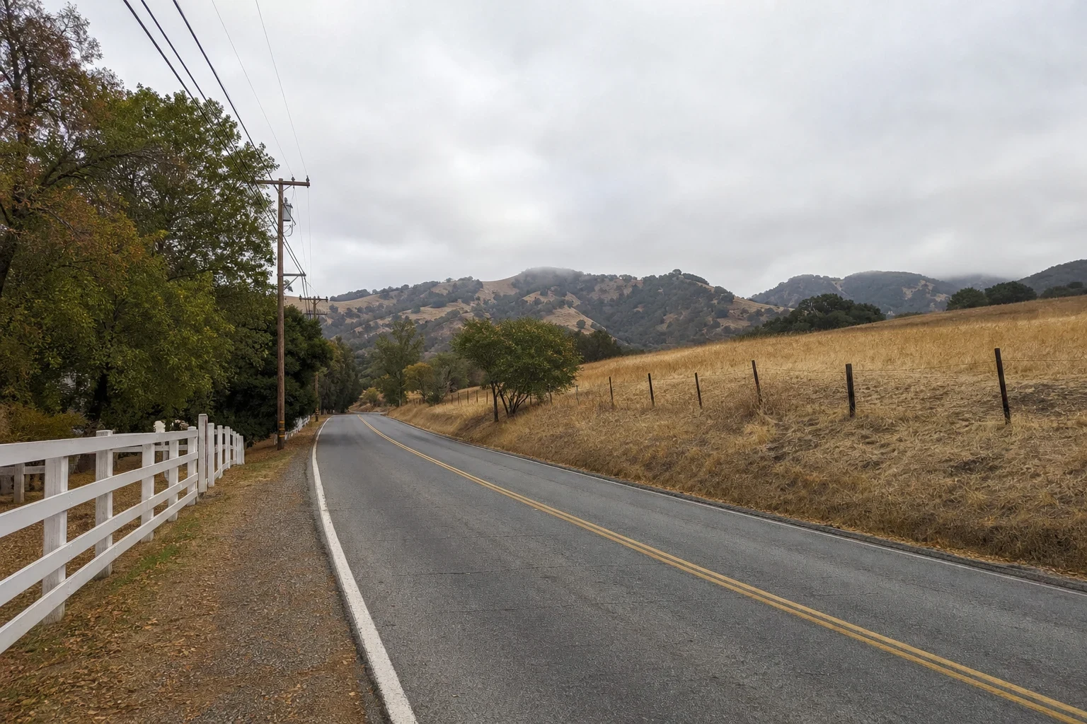 Quiet two-lane road on the edge of town with a white rail fence, utility poles, dry golden grass and oak-dotted hills under an overcast sky