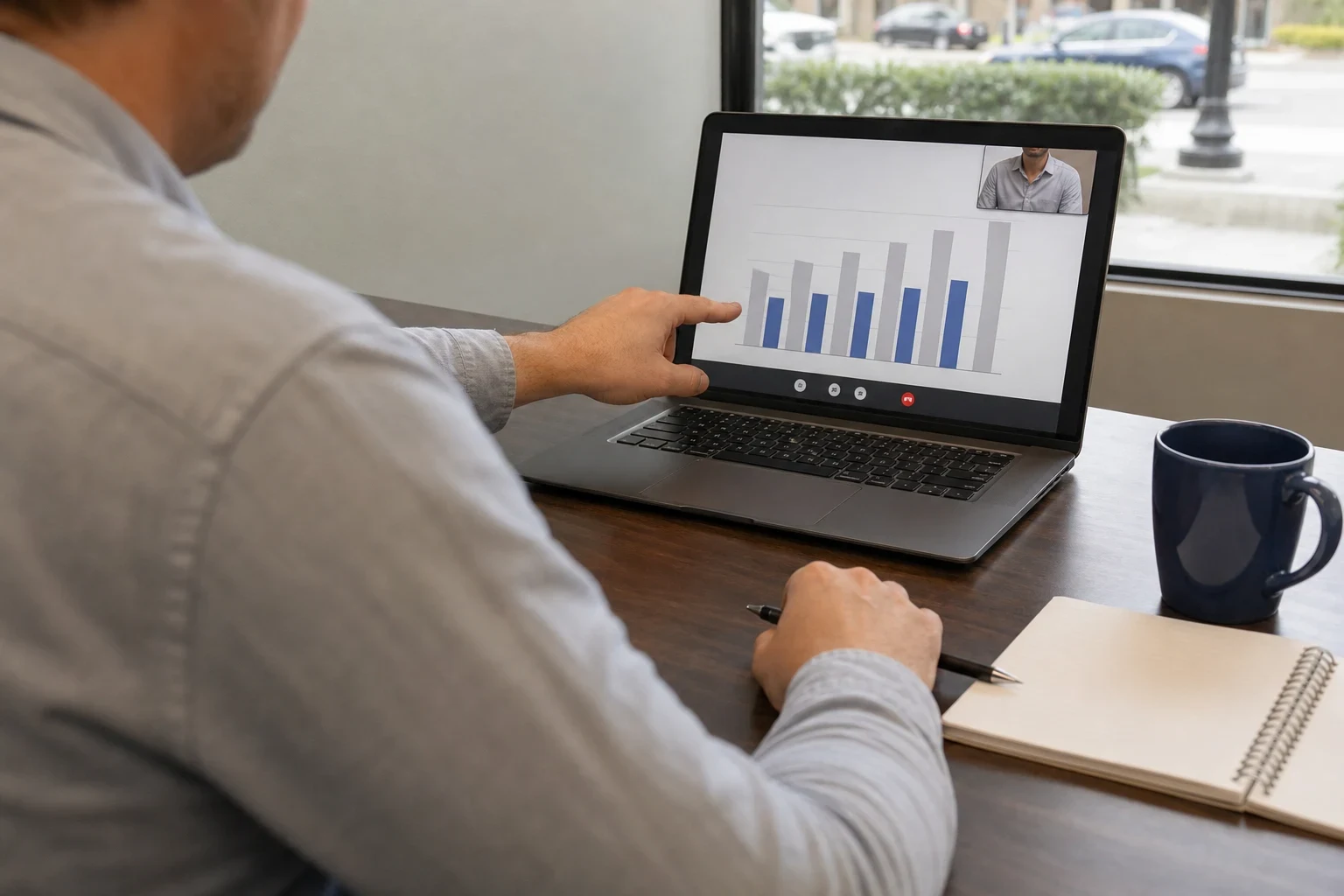 Man in a gray shirt at a wooden office desk pointing at a bar chart on a laptop during a video call, with a notebook, pen and navy mug nearby