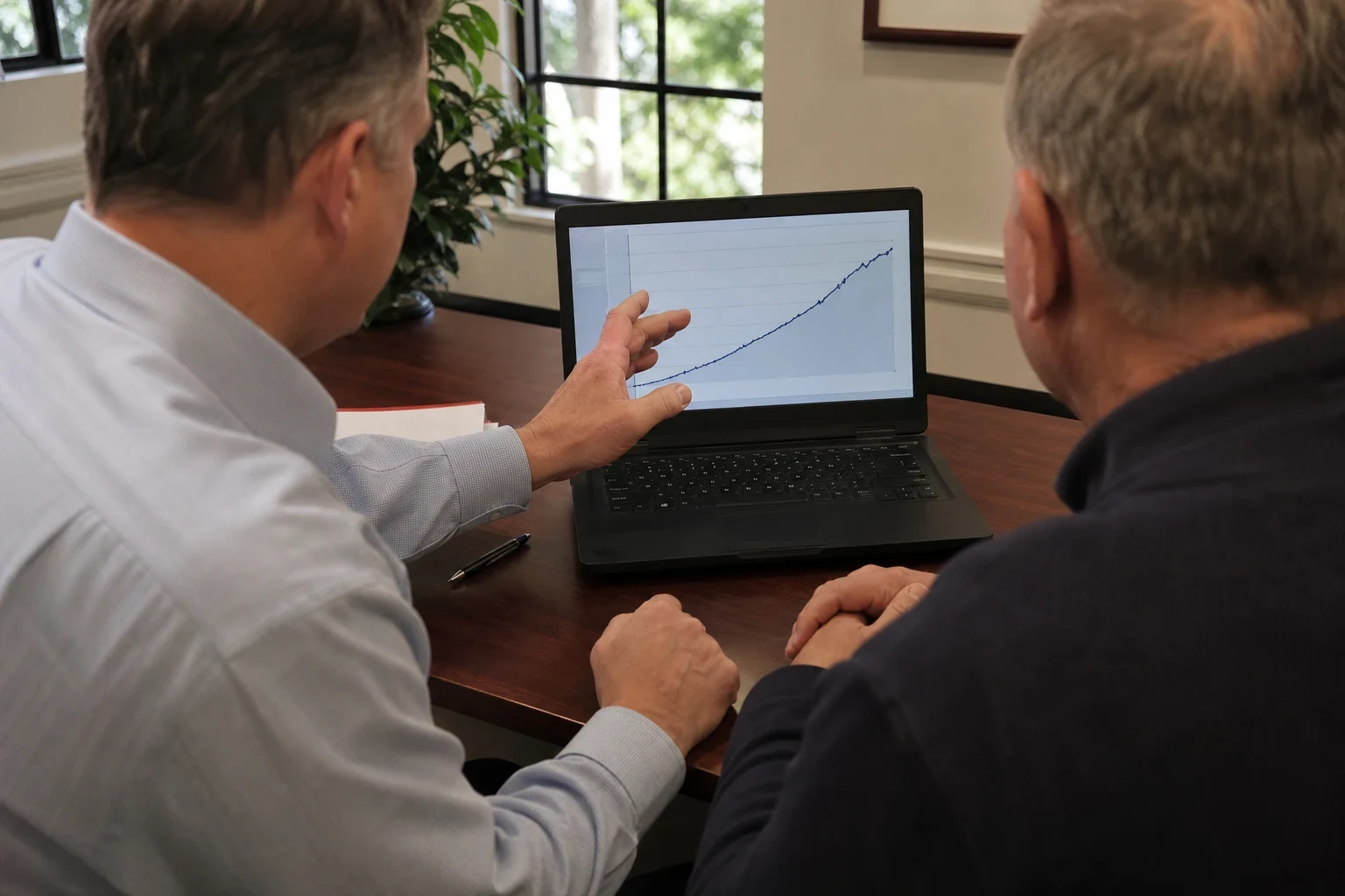 Agent in a light blue shirt points to a rising line chart on a laptop while an older client in a navy sweater looks on at an office desk by a window